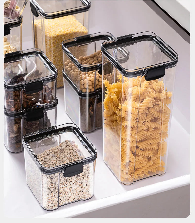 Clear storage containers with black lids holding various dry goods on a white surface.